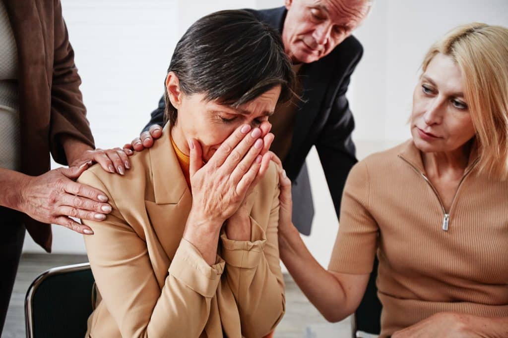 Mature woman with short hair sitting on chair and crying while people supporting her during psychotherapy session